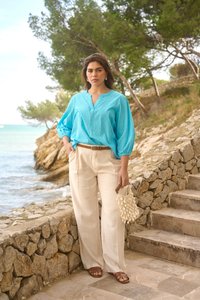 Femme debout près d'un mur en pierre et d'escaliers près de la mer, portant un chemisier bleu clair, un pantalon large crème, des sandales marron, tenant un sac en coquillages.