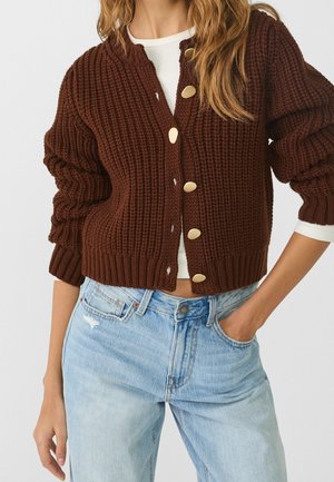 Femme portant un cardigan en tricot épais marron boutonné par-dessus un haut blanc et un jean taille haute bleu clair avec une main dans la poche.