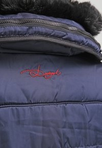 Interior de chaqueta acolchada en color navy con borde de piel sintética negra en el cuello, presenta un logo bordado en rojo y una línea de cremallera visible.