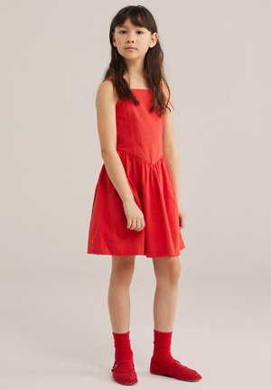 Fată tânără în picioare, purtând o rochie roșie fără mâneci, șosete roșii și pantofi roșii împletiți, pe un fundal simplu și neutru.