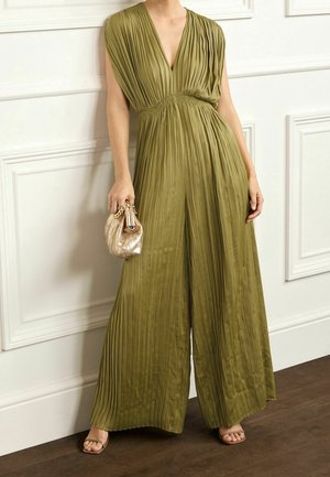 Femme portant une combinaison pantalon plissée kaki à jambes larges, tenant un petit sac à main métal doré, debout sur un sol en bois foncé devant un mur blanc à panneaux.