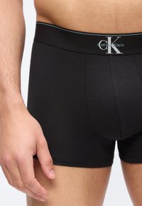 Κοντινό πλάνο στον κορμό και το πάνω μέρος του μηρού ενός άνδρα που φοράει μαύρα ανδρικά εσώρουχα Calvin Klein με λογότυπο στη μέση, το χέρι χαλαρό στο πλάι.