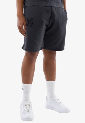 Persona che indossa pantaloncini neri con un logo in rilievo sulla coscia sinistra, calzini bianchi e scarpe da ginnastica bianche, in piedi su uno sfondo uniforme.