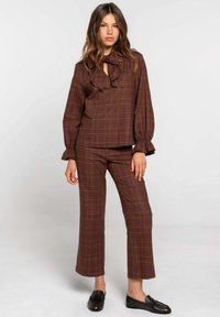 Mujer de pie vestida con una blusa marrón de cuadros y un lazo en el cuello, junto con pantalones a juego, combinados con mocasines negros, contra un fondo blanco liso.