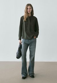 Camicia verde scuro a maniche lunghe con dettagli plissettati e bottoni, abbinata a jeans a zampa chiari. Borsa nera in mano. Sfondo neutro.