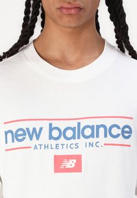 Hvit bomull T-skjorte med blå "new balance" tekst og "ATHLETICS INC." i rødt. Inkluderer en liten rød firkantlogo med "NB" under.
