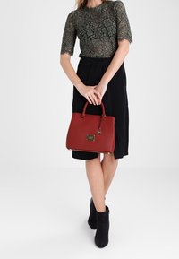 Röd texturerad handväska med dubbla handtag, guldfärgade detaljer och ett varumärkesplakett. Enkel, strukturerad design med en slät yta.