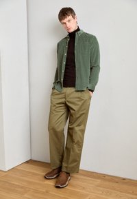Camasa verde din căptușeală, pulover negru cu guler înalt, pantaloni khaki și pantofi roz din piele maro. Camasa are un buzunar și nasturi din alamă.