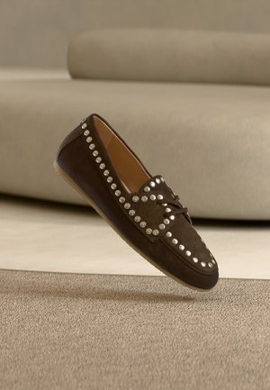 Καφέ μοκασίνια από σουέντ με ασημένια καρφιά κατά μήκος των πλευρών, στρογγυλεμένη μύτη και φιόγκο μπροστά. Επίπεδη σόλα με ελάχιστη υφή.