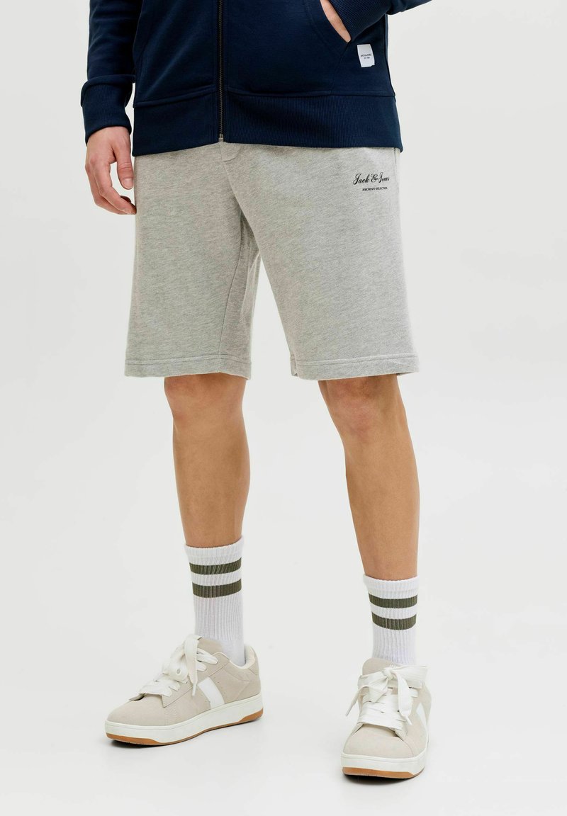 Mann trägt graue Jack & Jones Shorts, weiße Socken mit zwei dunkelgrünen Streifen, hellbeige Sneaker und eine marineblaue Zip-up-Jacke.