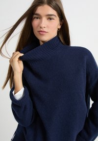 Granatowy sweter dziany z grubym ściągaczem w kołnierzu i szerokimi rękawami, o miękkiej fakturze i luźnym kroju.