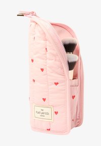 Rosa quiltad sminkborsthållare med röda hjärtan, sidokudde, och två borstar synliga inuti. Etikett på framsidan: "THE FLAT LAY CO. LONDON."