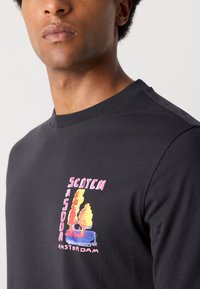 Sort bomulds langærmet t-shirt med et farverigt grafisk print af træer og teksten "SCOTCH AMSTERDAM" på venstre side.