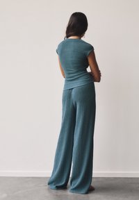 Teal ribbet strikbluse med korte ærmer, parret med lyse teal breddebukser. Blød tekstur og afslappet pasform, set fra ryggen.