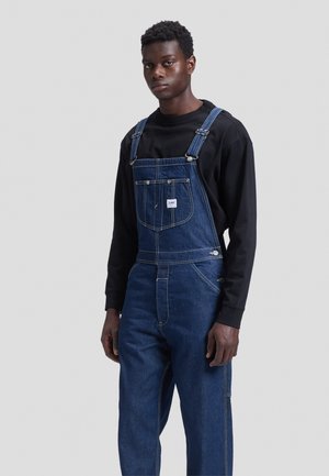 Jeune homme portant une salopette en jean bleu foncé et une chemise noire à manches longues, debout devant un fond gris clair uni.