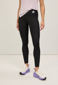Leggings neri con vita alta, dotati di un logo bianco. Abbinati a sneakers chiare con accenti viola. Texture liscia.