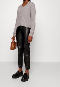 Žena v šedém pleteném svetru, černých kožených kalhotách, černých mohutných mokasínech a držící malou hnědou kabelku na jednobarevném pozadí.