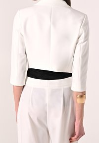 Witte, op maat gemaakte jas met driekwart mouwen, een zwarte tailleband en glanzende gouden manchetdetails, met een gestructureerd en modern ontwerp.