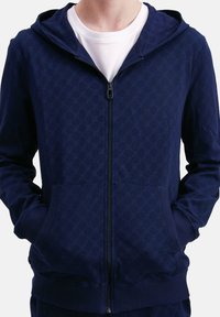 Persona che indossa una felpa con zip blu navy con un leggero motivo geometrico e una t-shirt bianca a girocollo sotto. Mani nelle tasche frontali.