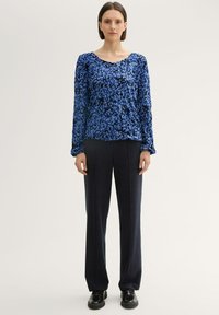 Blaue, blumenbedruckte Langarmbluse mit Rundhalsausschnitt, kombiniert mit dunklen, geraden Hosen und schwarzen Schuhen, getragen von einem Model vor einem hellen Hintergrund.