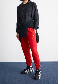 Svart långärmad tröja med texturerat mönster, röda sweatpants och svarta sneakers med grå detaljer och vit branding, allt mot en neutral bakgrund.