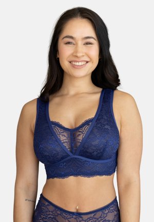 Woman smiling wearing matching dark blue lace bralette and high-waisted lace underwear against a plain background.