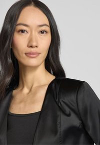 Blazer in satin nero con un design strutturato, caratterizzato da un collo rotondo e una texture morbida e liscia, indossato sopra un top nero senza maniche.