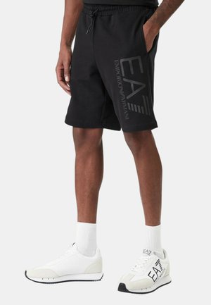 Uomo che indossa pantaloncini neri Emporio Armani EA7, calzini bianchi EA7 e sneakers bianche, in piedi con le mani in tasca.