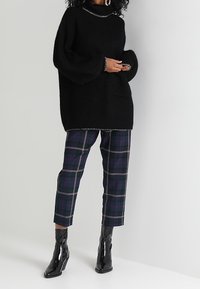 Czarny dziany sweter z wysokim kołnierzem i przednią kieszenią, zestawiony z kraciastymi spodniami w ciemnoniebieskie, zielone i bordowe wzory oraz błyszczącymi czarnymi butami.