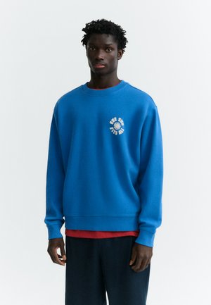 Homme à la peau foncée et aux cheveux bouclés portant un sweat bleu avec un texte circulaire blanc sur la poitrine, un sous-vêtement rouge et un pantalon foncé, debout devant un fond blanc.