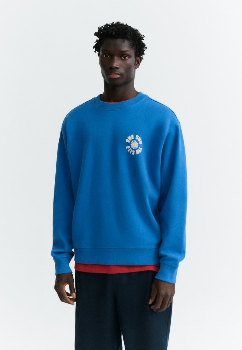 Homme à la peau foncée et aux cheveux bouclés portant un sweat bleu avec un texte circulaire blanc sur la poitrine, un sous-vêtement rouge et un pantalon foncé, debout devant un fond blanc.