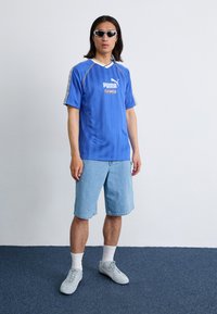 Modrá pruhovaná dres Puma s krátkymi rukávmi, s bielym golierom a logom; nosený s svetlými džínsovými šortkami a šedými teniskami.
