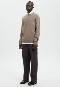 Beiger Wollpullover mit rundem Ausschnitt, kombiniert mit einem weißen Hemd mit Kragen, dunkler Hose und schwarzen Lederschuhen.