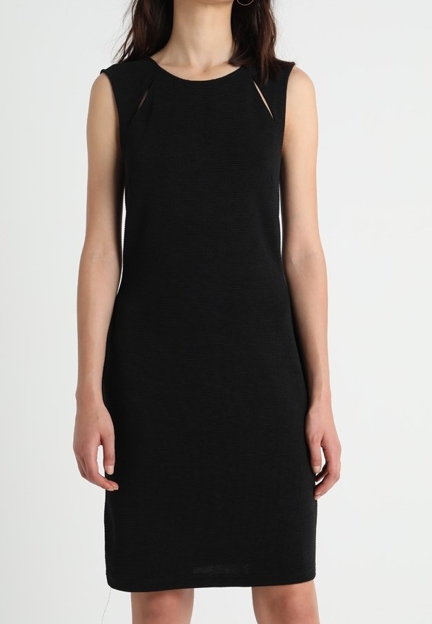Femme portant une robe noire sans manches à la longueur genou avec des détails subtils d’ouverture aux épaules, debout devant un fond blanc uni.