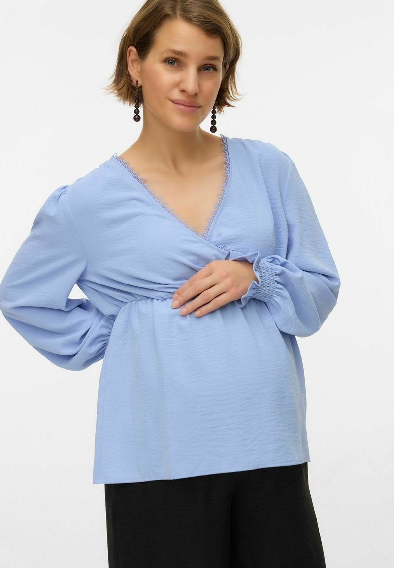 Lyseblå graviditetst-shirt med V-hals, rynket talje og puffede ærmer, med et fint bånd langs halsudskæringen. Bruges med sorte bukser.