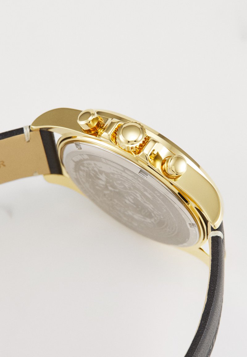 Orologio dorato con un cinturino in pelle nera testurizzata, quadrante argenteo con motivi intricati e tre pulsanti sul lato.