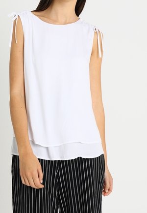 Woman wearing sleeveless white blouse with small shoulder ties and black pants with white vertical stripes, standing against a plain background.