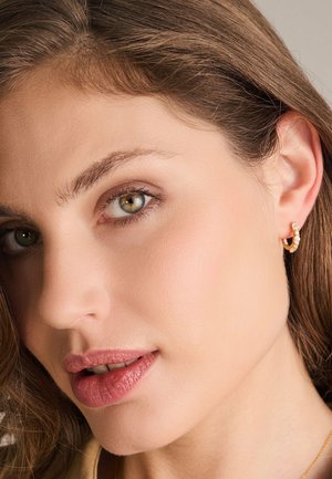 Guldörhängen i form av ringar med små pärlprydnader. Modellen har ljusbrunt hår, gröna ögon och en naturlig sminkning.
