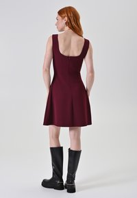 Bordeaux mouwloos jurkje met een lage rug en ritsdetails, gecombineerd met knie-hoge zwarte leren laarzen met gouden accenten.