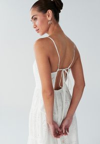 Robe blanche sans manches avec un dos échancré, des liens ajustables et une texture en broderie ajourée. Design fluide à volants superposés.