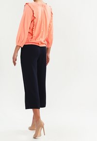 Korálový pletený top s volánkovým lemem, zapínání na knoflíky vzadu; v kombinaci s námořnicky modrými zkrácenými kalhotami a nahými lodičkami, hladká textura.