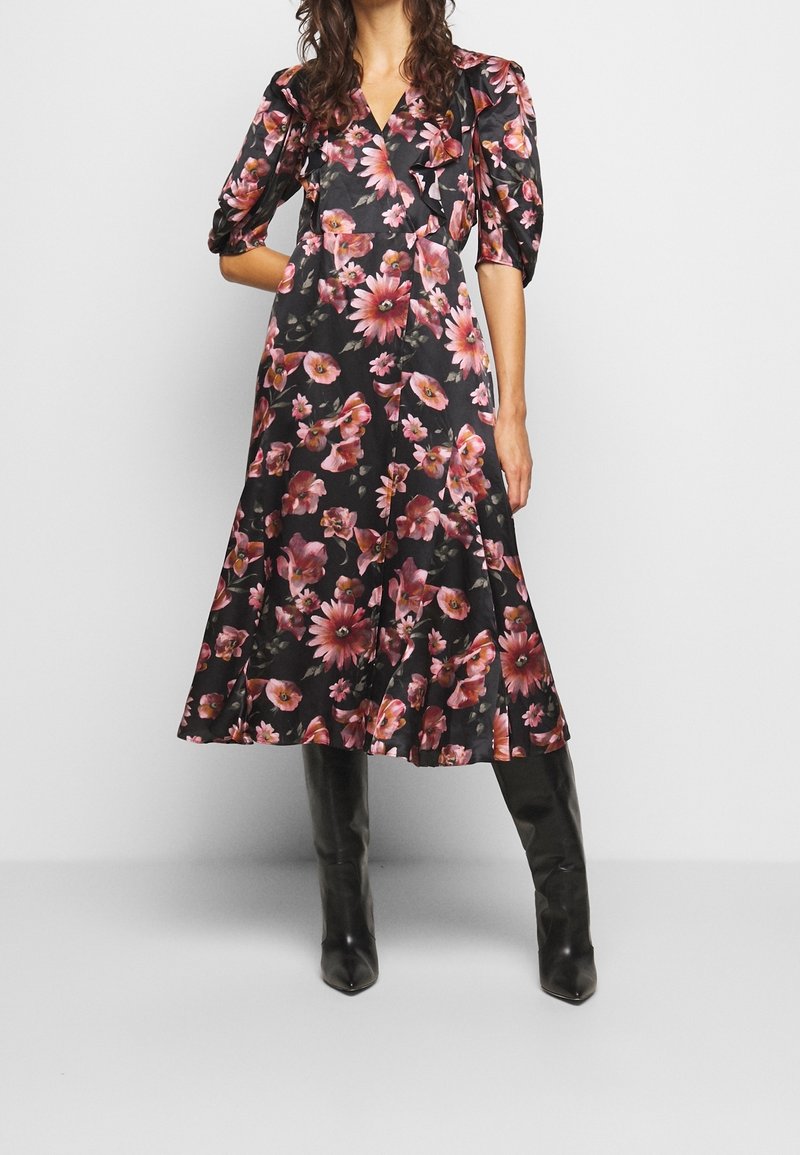 Femme portant une robe midi noire à imprimé floral rose, manches bouffantes et bottes noires montantes, debout devant un fond clair uni.