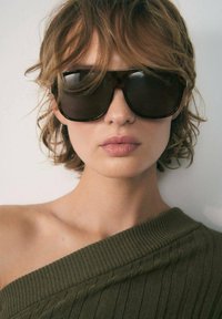Ochelari de soare oversized cu lentile întunecate, un cadru gros din torsaduș maro și o finisare netedă; purtați cu o bluză verde măslin cu umerii descoperiți.