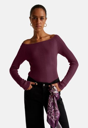Top en maille côtelée, décolleté bardot, à manches longues de couleur violet profond, associé à un jean noir taille haute. Une écharpe violette à motifs est nouée à la taille.
