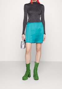 Glitzerndes schwarzes Langarmoberteil, türkiser Minirock, grüne Wildleder-Plateau-Stiefeletten und eine kleine silberne Handtasche. Glatte Texturen und lebendige Farben.