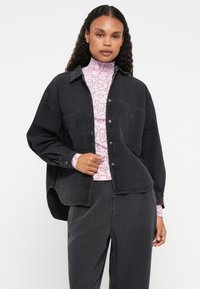 Čierna oversized denimová košeľa s dvoma náprsníkovými vreckami, gombíkovým zapínaním a zakriveným lemom, nosená cez ružový bodkovaný rolák.