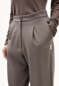 Grijze geplooide broek met een gladde textuur, voorzien van een knoopsluiting en zijzakken, aangevuld met een klein merkenlabel aan de zijkant.