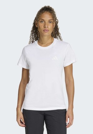 Kvinde med krøllet hår iført en enkel hvid kortærmet trøje og sorte bukser, stående mod en lys baggrund.