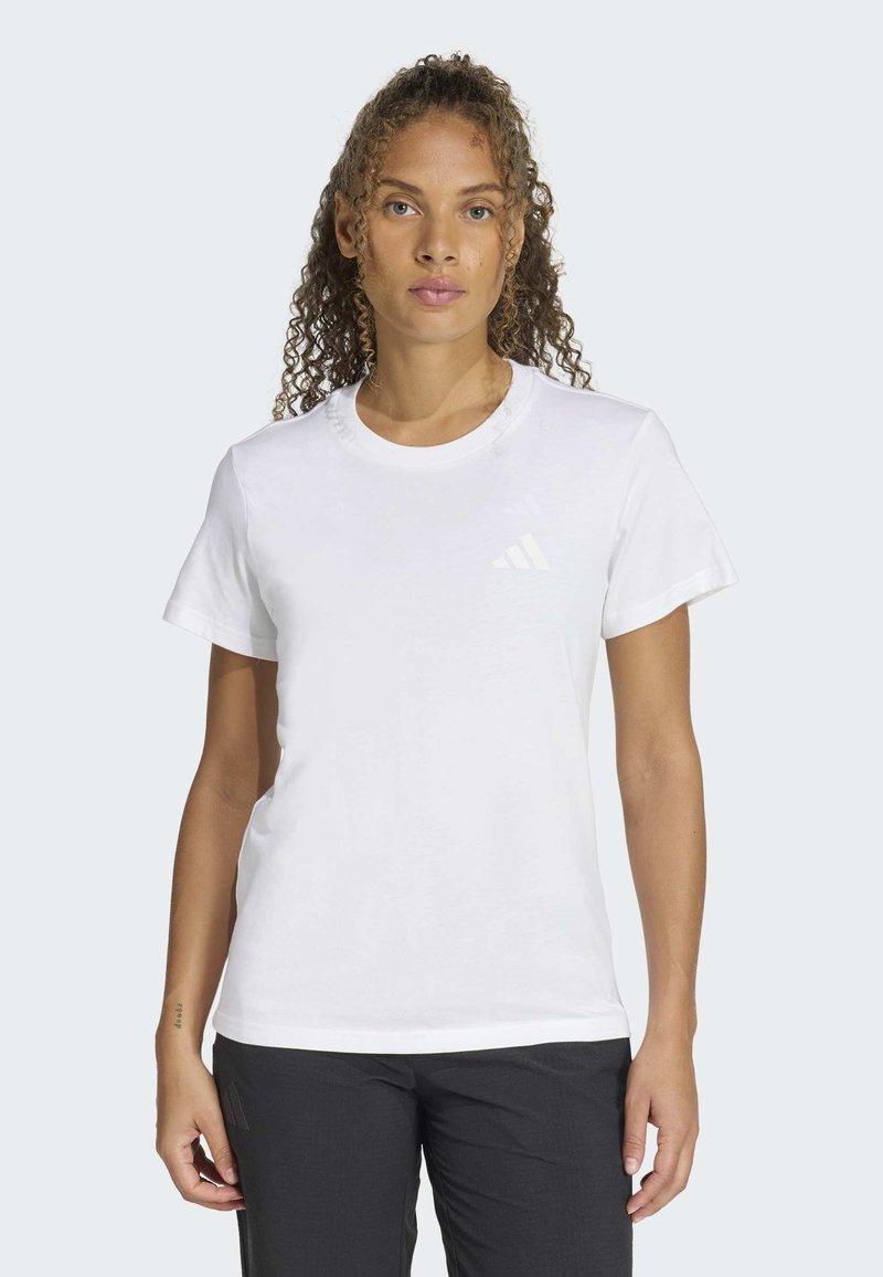 Žena s kudrnatými vlasy, která má na sobě jednoduché bílé tričko s krátkým rukávem a černé kalhoty, stojící před světlým pozadím.