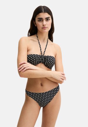 Kobieta z długimi ciemnymi włosami, ubrana w czarne bikini z białymi wzorami żaglówek, z założonymi skrzyżowanymi ramionami, stojąca na jednolitym jasnym tle.